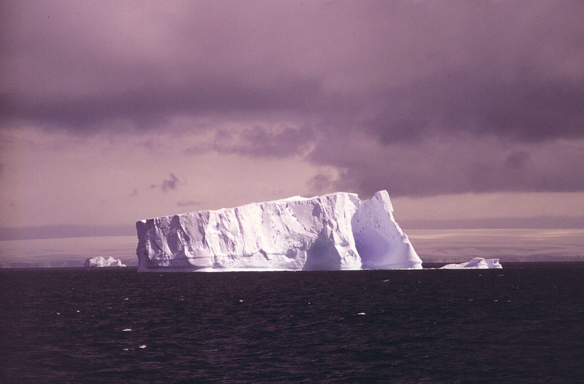 A huge flat-topped iceberg with sheer white sides on a dark sea under heavy purple-grey cloud, smaller bergs on the horizon