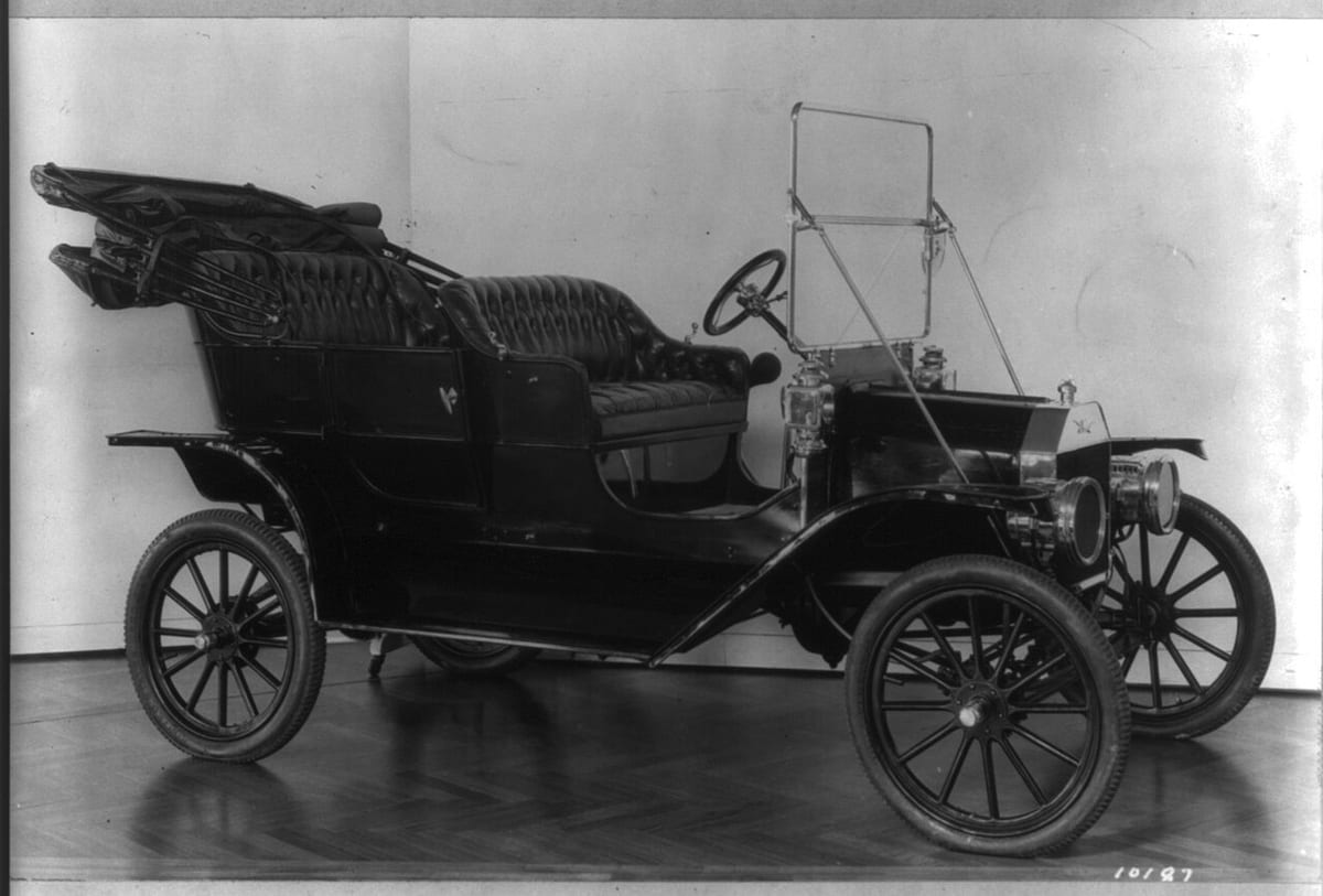 Black-and-white photo of a black open-top Ford Model T touring car with a folded hood, tufted leather seats, brass lamps and spoked wheels