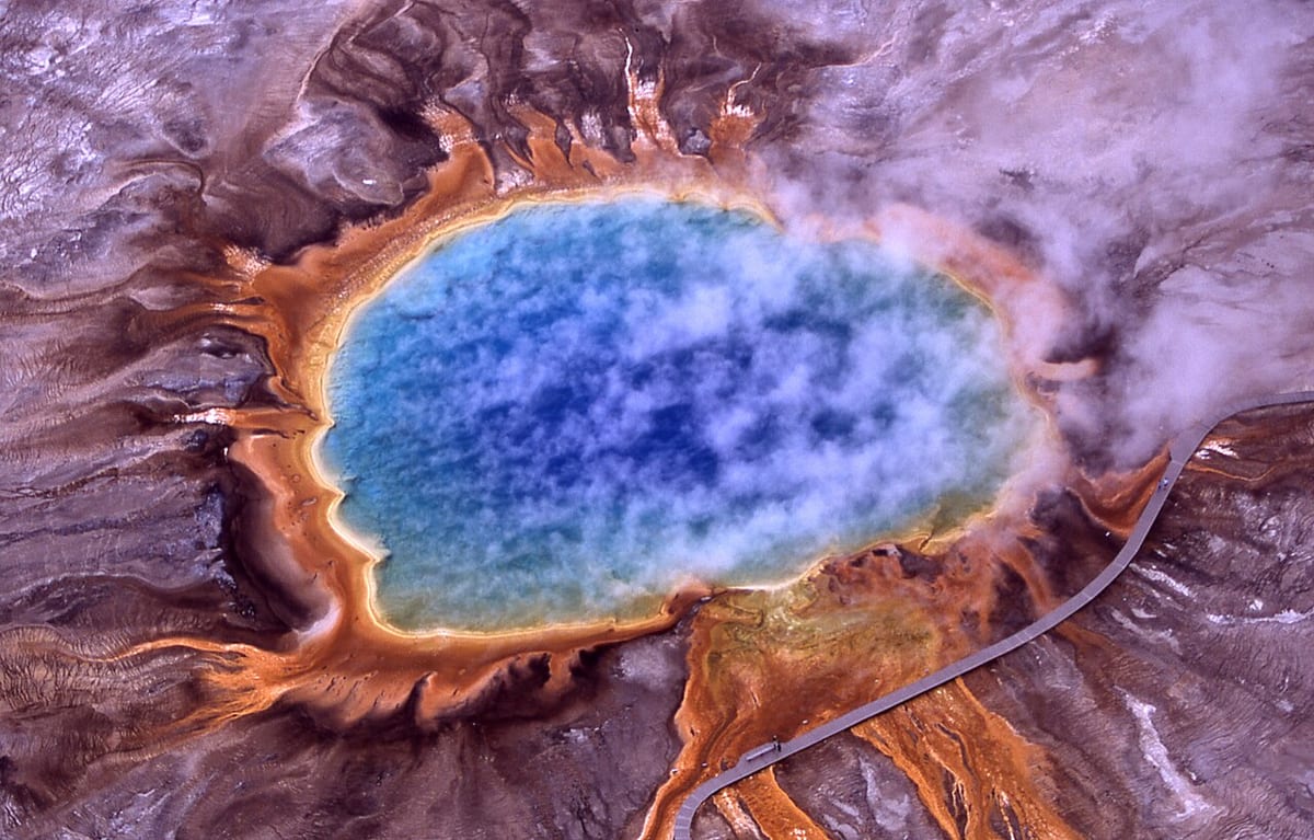 Aerial view of a vast hot spring: steaming deep-blue water ringed by yellow and bright orange bands that streak out across brown ground, a boardwalk beside it