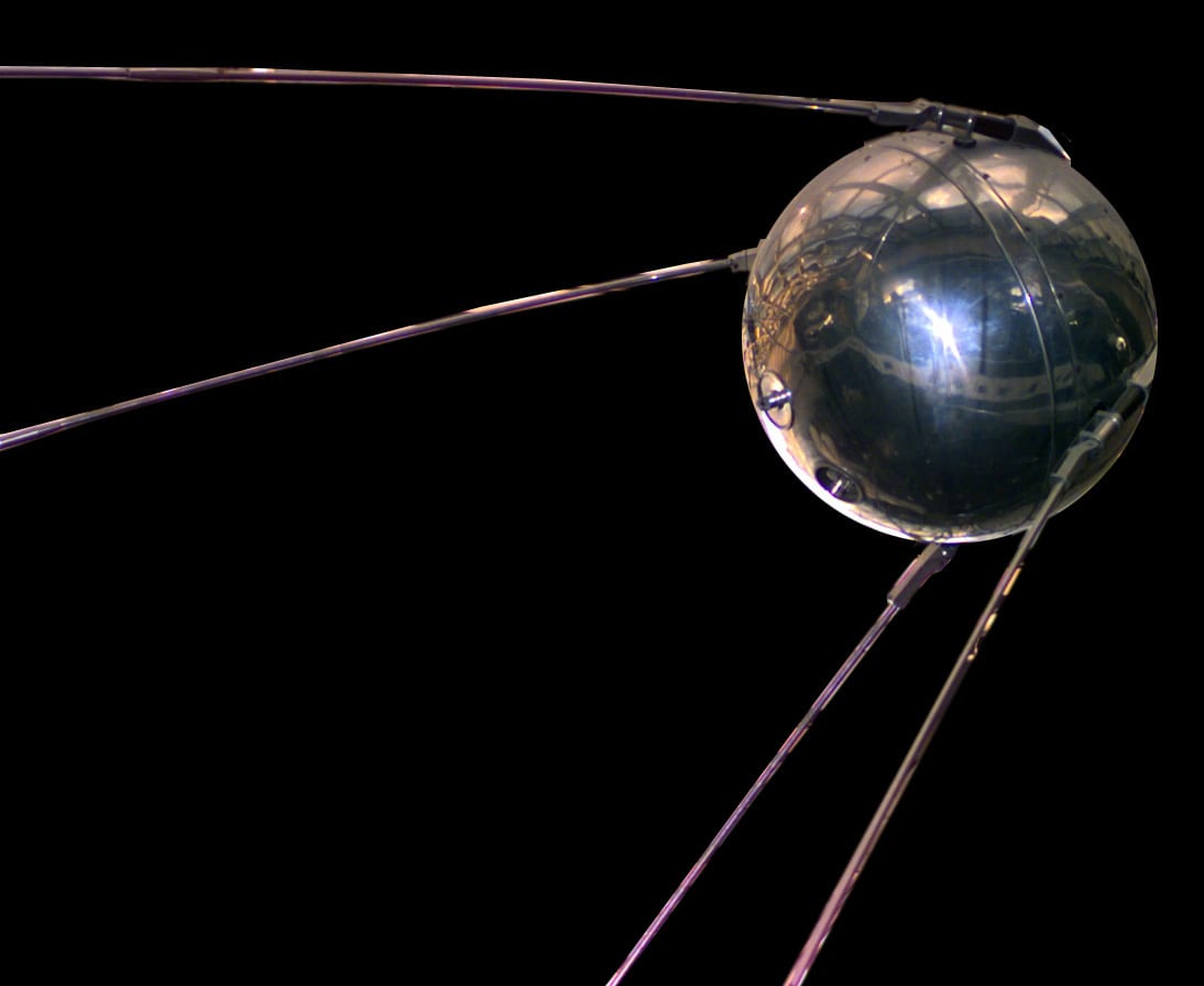 A polished metal sphere with four long rod antennas sweeping back from it, against a black background
