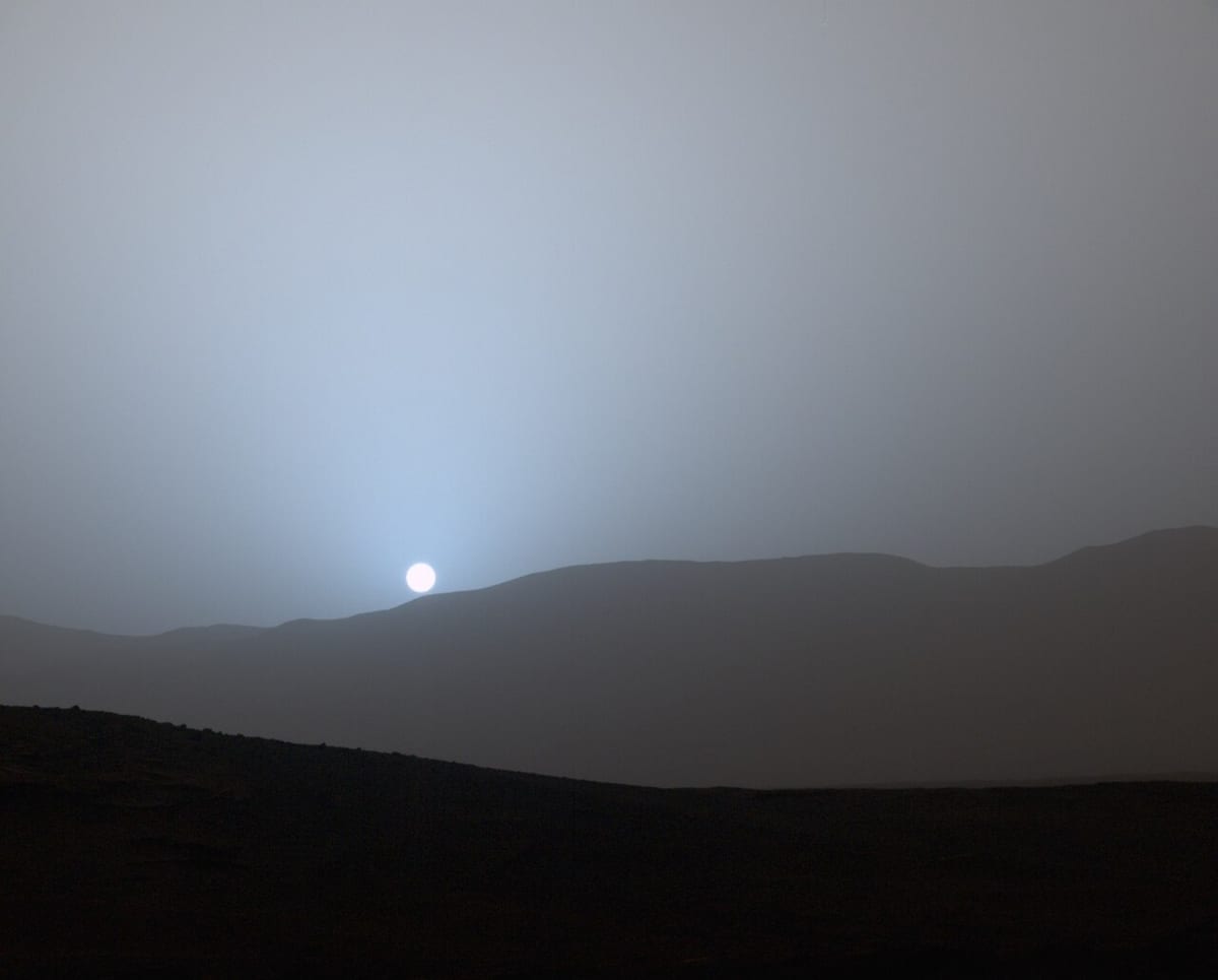 A small white sun setting over low dark hills on Mars, a blue glow around it in an otherwise dusty grey sky