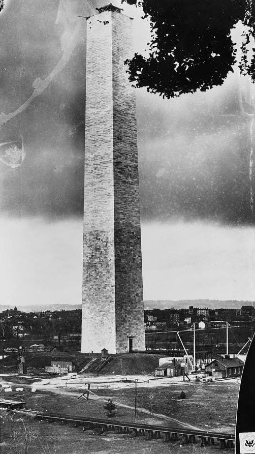 Black-and-white photo of a tall unfinished stone obelisk with lifting gear on its flat top, above work sheds and a rail track, a city behind