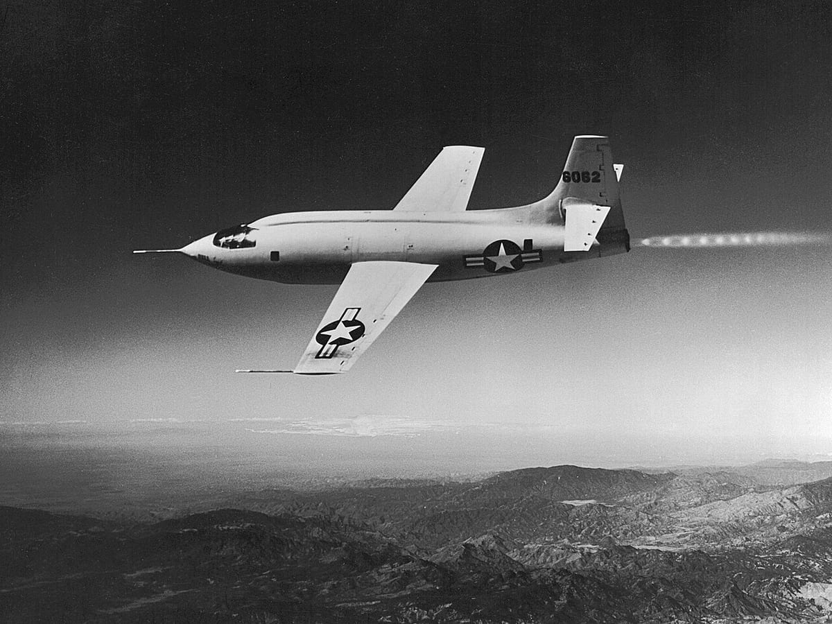 Black-and-white photo of a bullet-shaped rocket plane with straight wings and US star markings flying above mountains, flame trailing from its tail