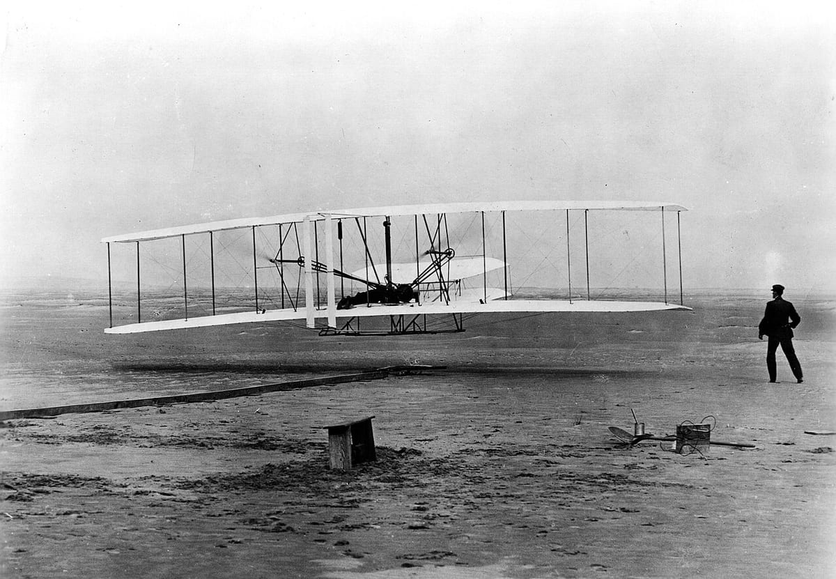 Black-and-white photo of the Wright Flyer, a white biplane, lifting a few feet off the sand with a man lying at the controls and another watching at right