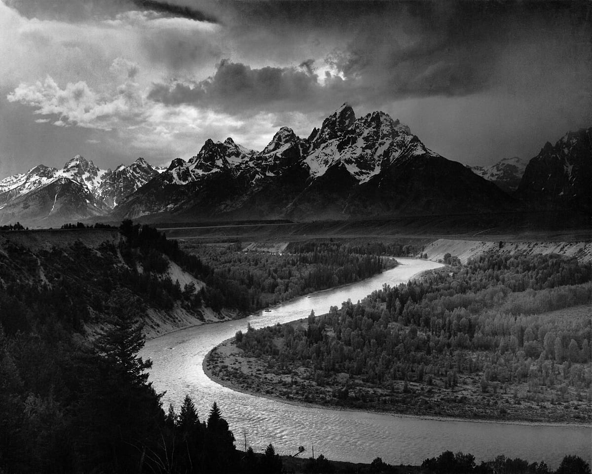 Black-and-white photo of the Snake River curving through dark forest below the jagged, snow-streaked Teton peaks under a stormy sky