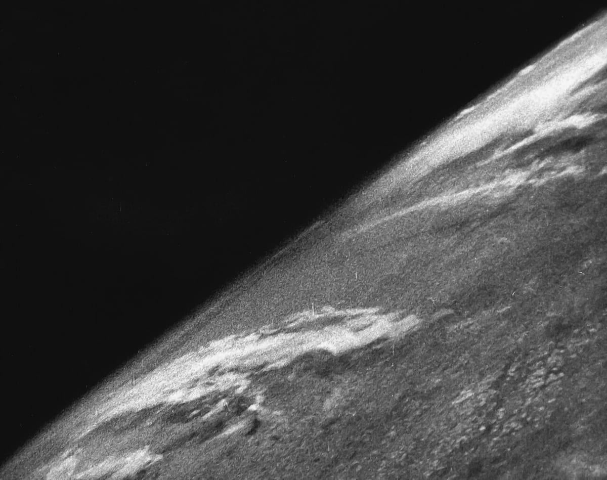 Grainy black-and-white photo of the curve of the Earth seen from high above, streaks of cloud over the ground and the black of space beyond