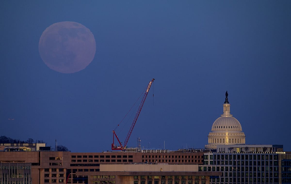 A pinkish full moon in a deep blue dusk sky over Washington, with the lit dome of the US Capitol and a red crane among the buildings below